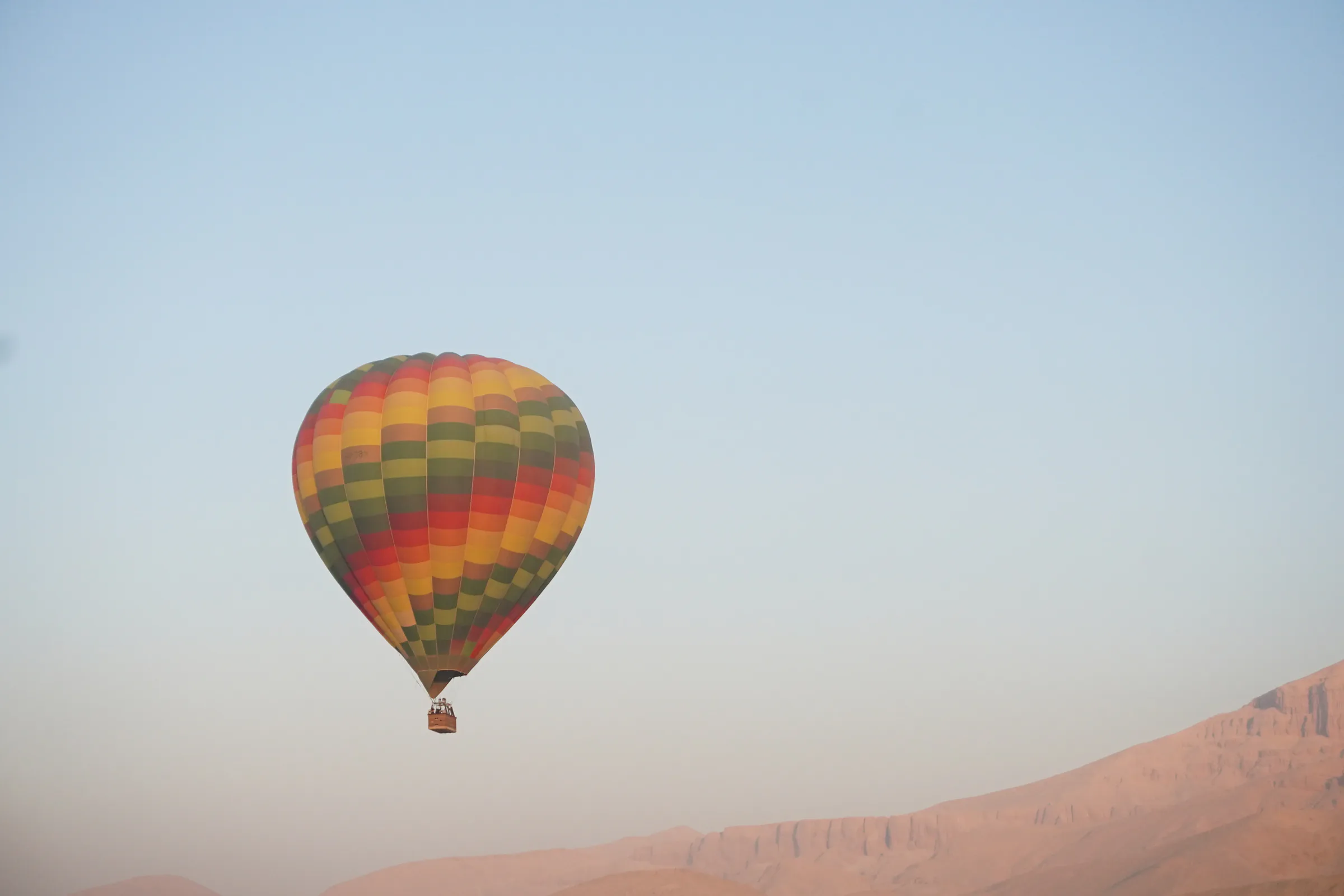 A red, orange and green checkered hot air balloon floating above hazy desert cliffs