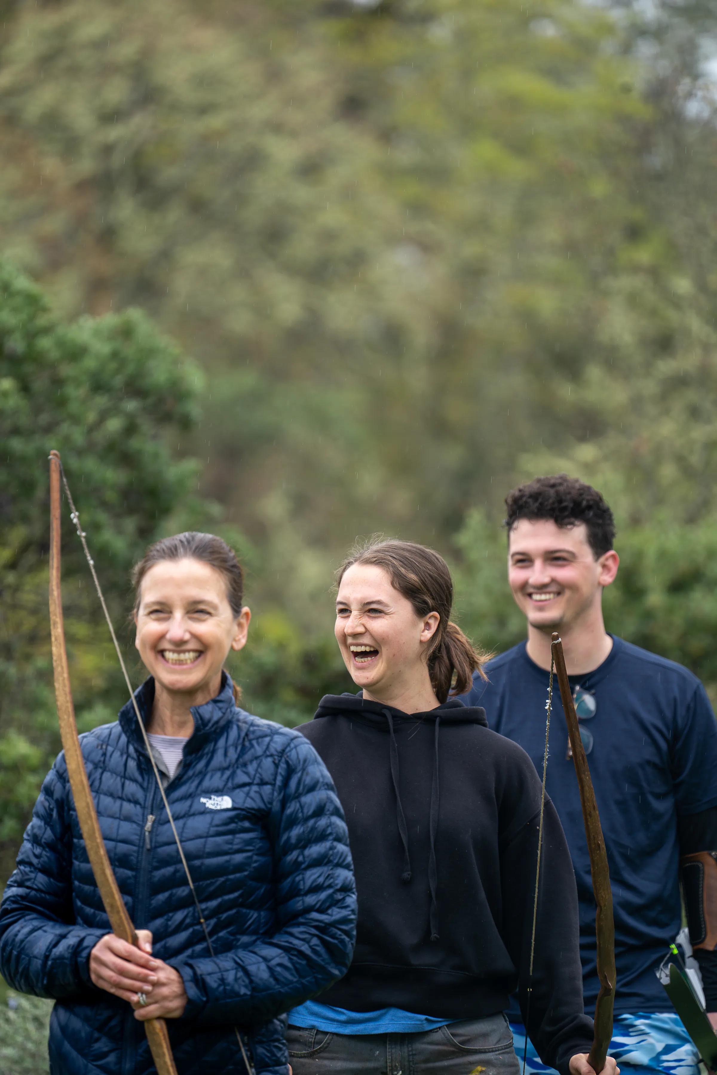 Three people holding longbows, laughing together on an overcast afternoon