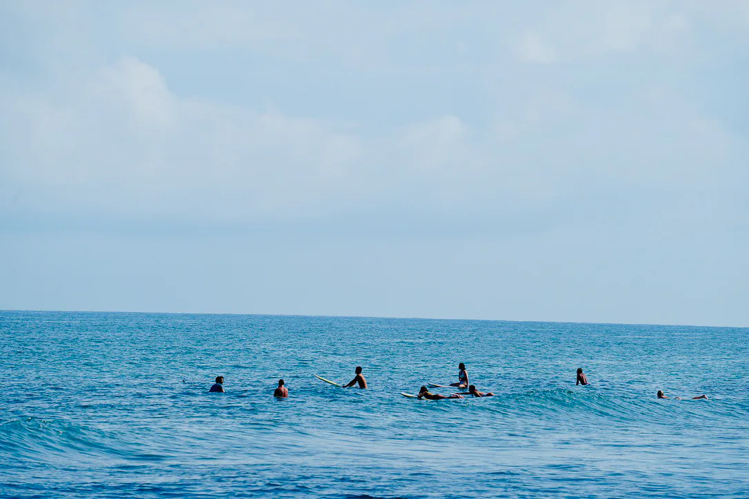 A line of surfers waiting beyond the waves under a pale blue sky