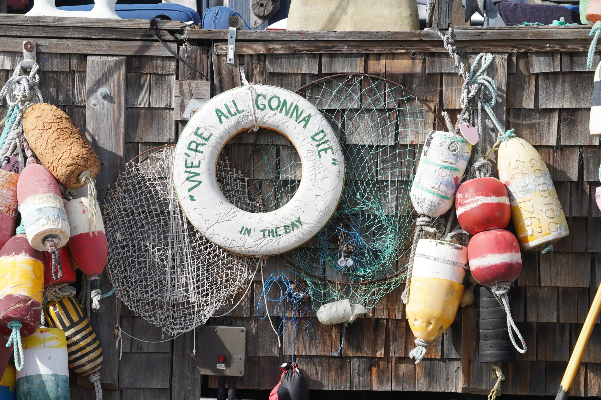 Weathered lobster buoys, netting and a joke life ring reading We are all gonna die in the bay, hanging on a shingle wall